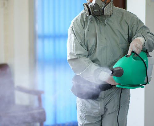 a person wearing a mask and gloves spraying a green container to remove odor
